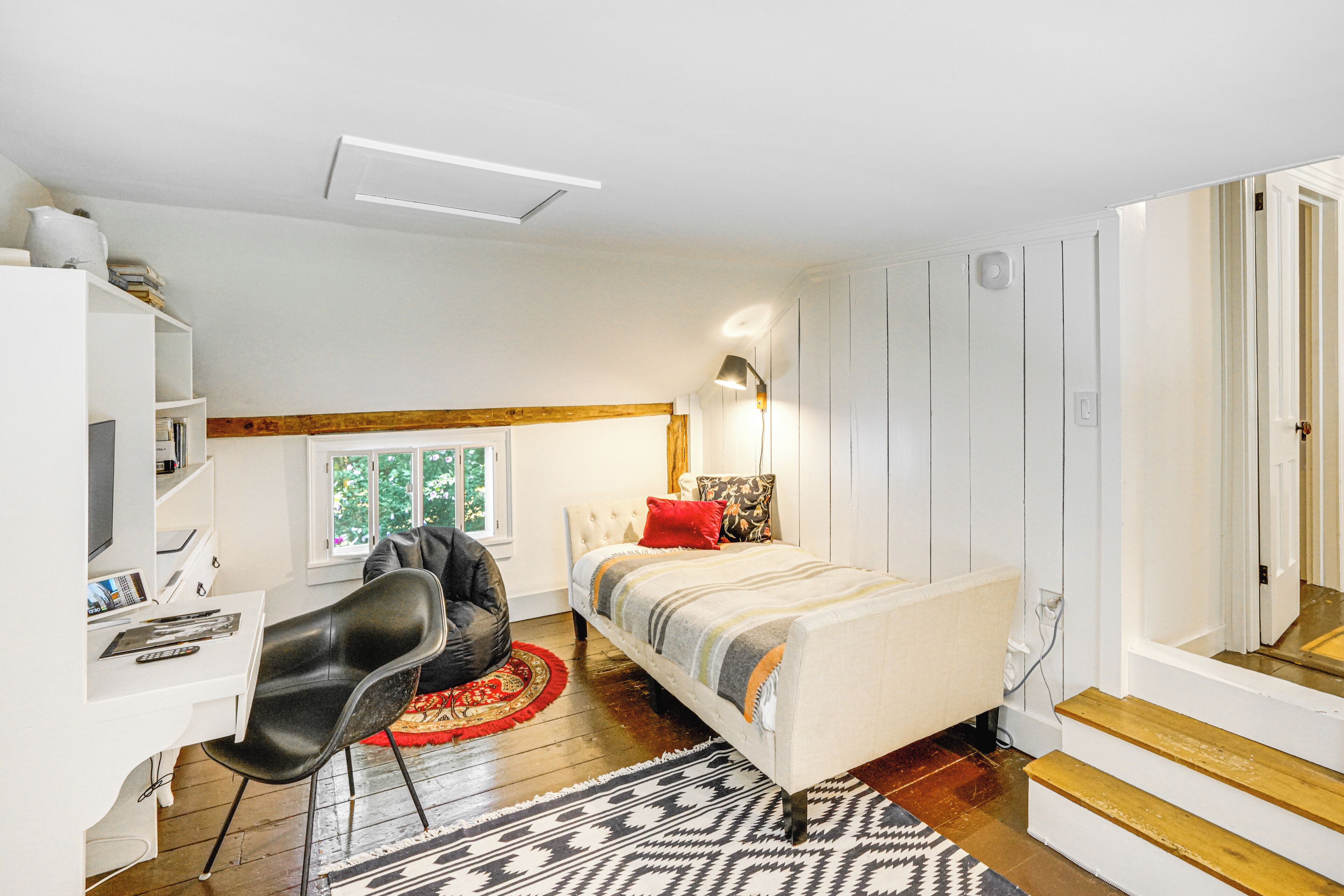A compact bedroom with a bed, a desk, and a chair. There is a window providing natural light, and a mirror is visible on the wall.