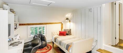 A compact bedroom with a bed, a desk, and a chair. There is a window providing natural light, and a mirror is visible on the wall.
