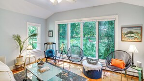 A spacious living room with large windows, a ceiling fan, and a variety of seating options including a chair with an orange cushion.