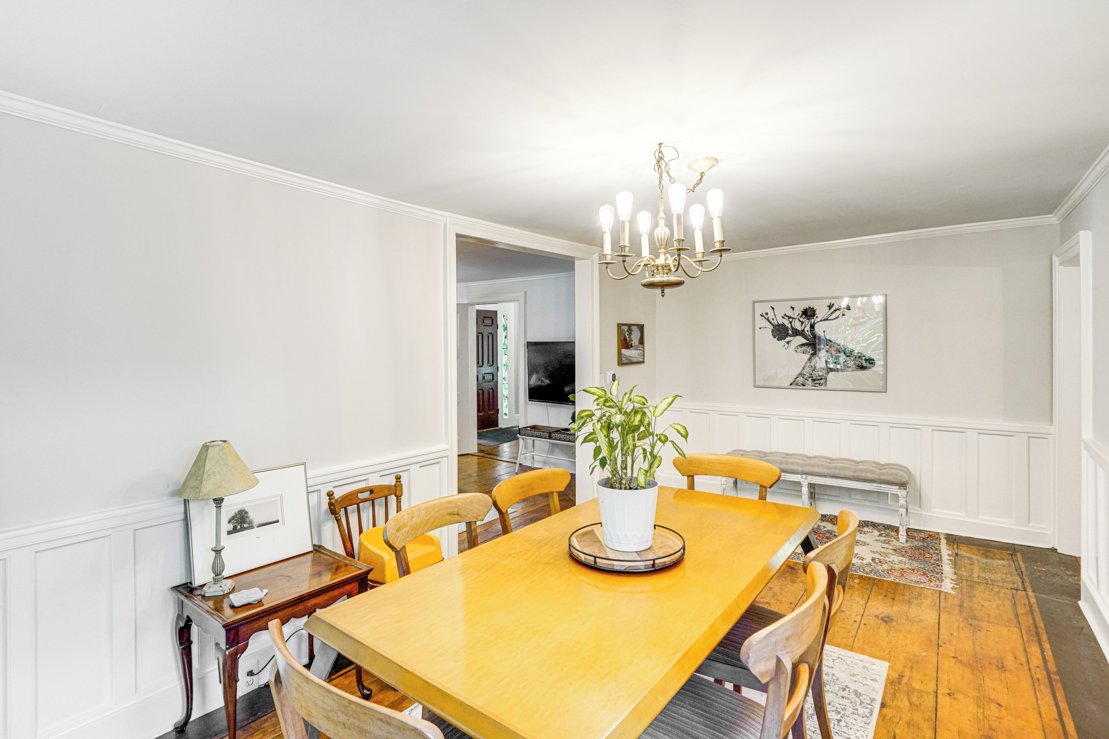 A dining room with a wooden table, chairs, a chandelier, a potted plant, and a painting on the wall.