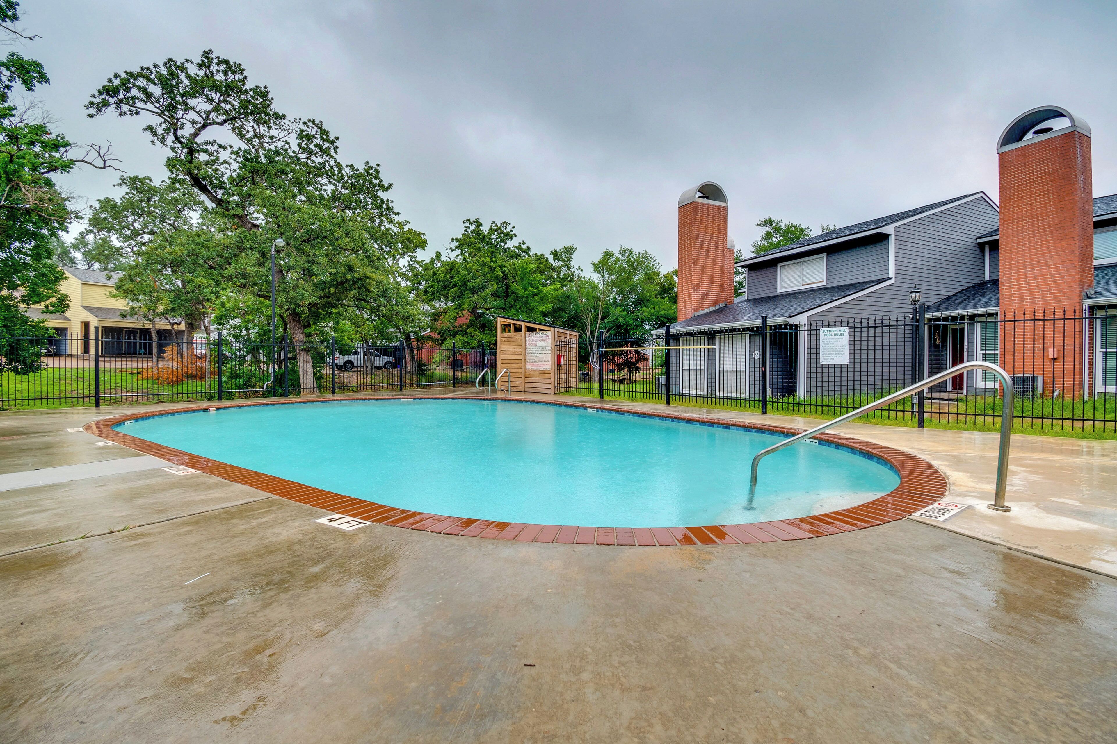 A swimming pool with a brick border, surrounded by a fence, and adjacent to a building with a brick chimney.