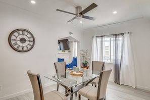 Uma sala de jantar moderna com mesa de vidro, quatro cadeiras, ventilador de teto, relógio de parede grande e vista para o exterior através de cortinas finas.