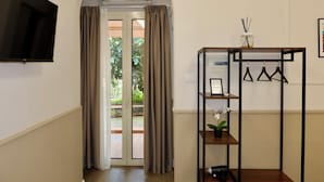 Ein Zimmer mit an der Wand montiertem Fernseher, einer Schiebeglastür mit Vorhängen, einem Holzregal mit Pflanze und einem kleinen Tisch mit Vase.
