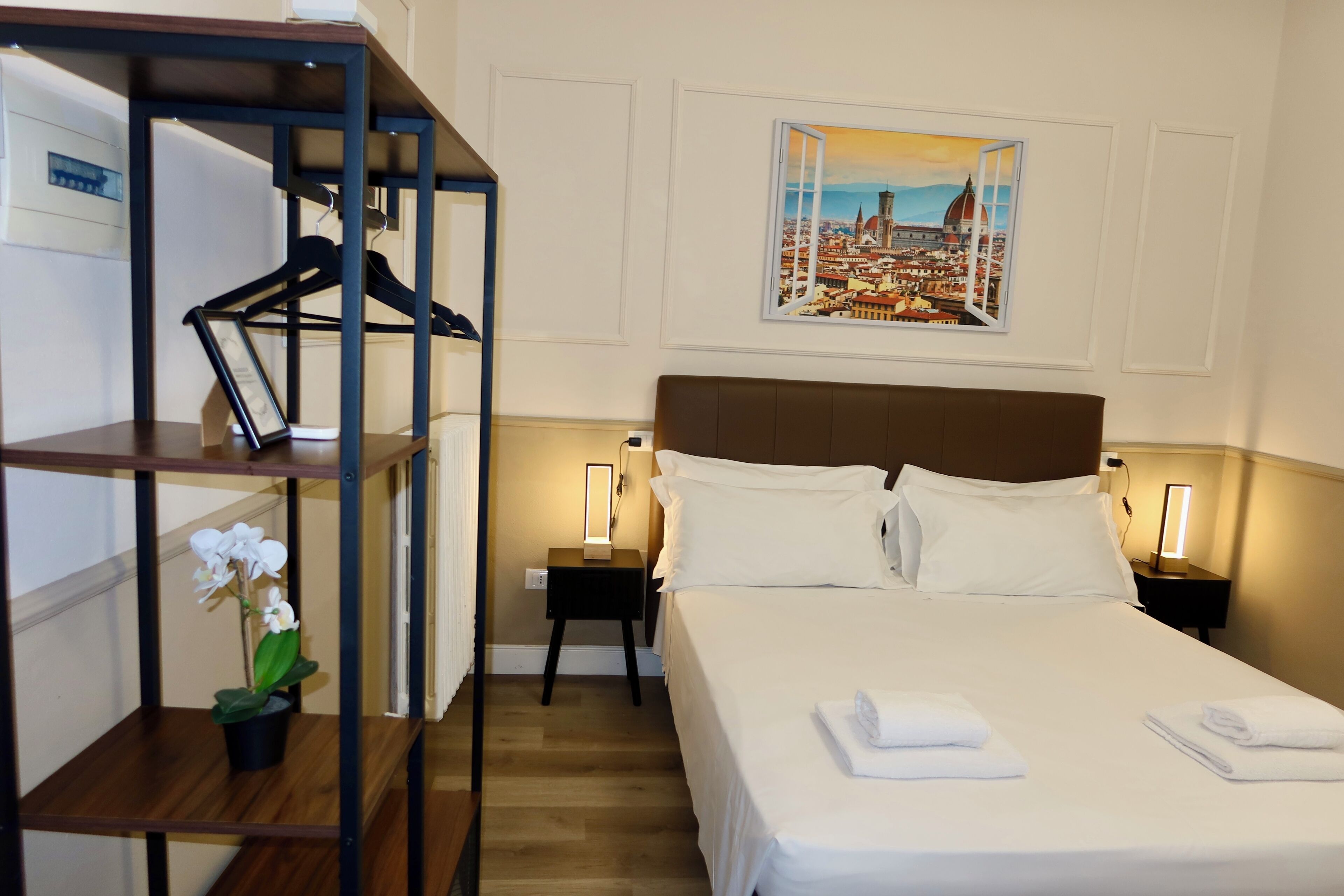Habitación de hotel con cama, mesita de noche, estante en la pared con una planta y un cuadro enmarcado de un paisaje urbano.