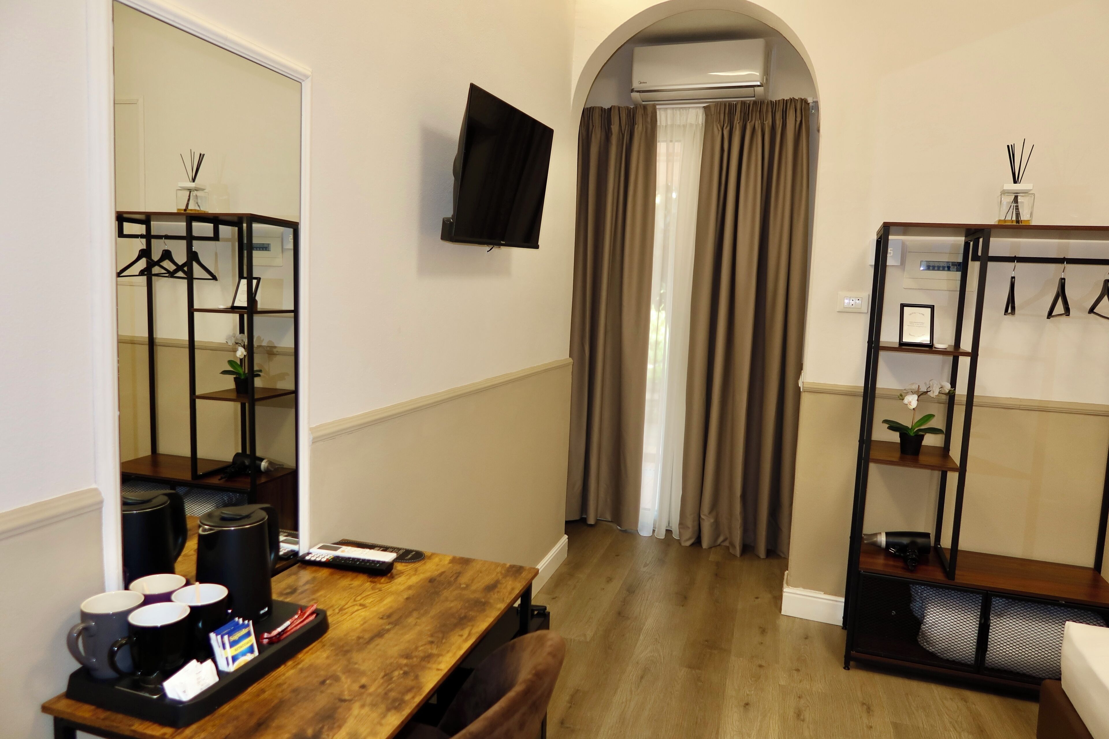 A hotel room with a wooden table, a TV mounted on the wall, a coat rack, and a window with curtains.