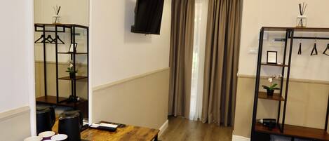A hotel room with a wooden table, a TV mounted on the wall, a coat rack, and a window with curtains.