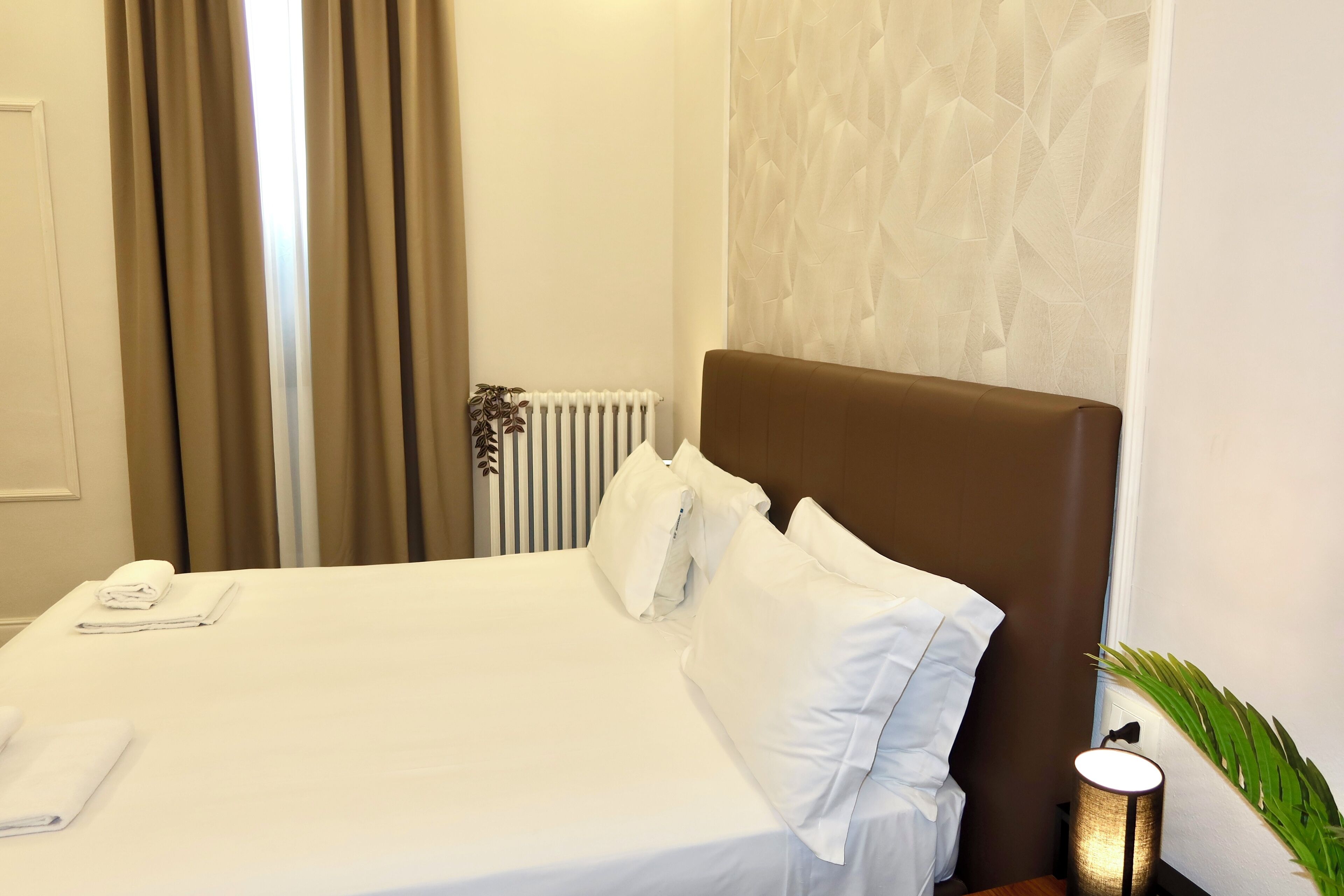 A neatly made bed with white linens and pillows, a radiator, a plant, and a lamp on a bedside table.