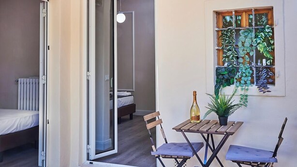 Ein Zimmer mit einem Bett, einem kleinen Tisch mit Flasche und Pflanze sowie einem Fenster mit hängenden Pflanzen.