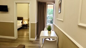 Ein Hotelzimmer mit Bett, Nachttisch, Lampe und einem Tisch mit einer Pflanze. Ein Fernseher ist an der Wand montiert und es gibt ein Fenster mit Vorhängen.
