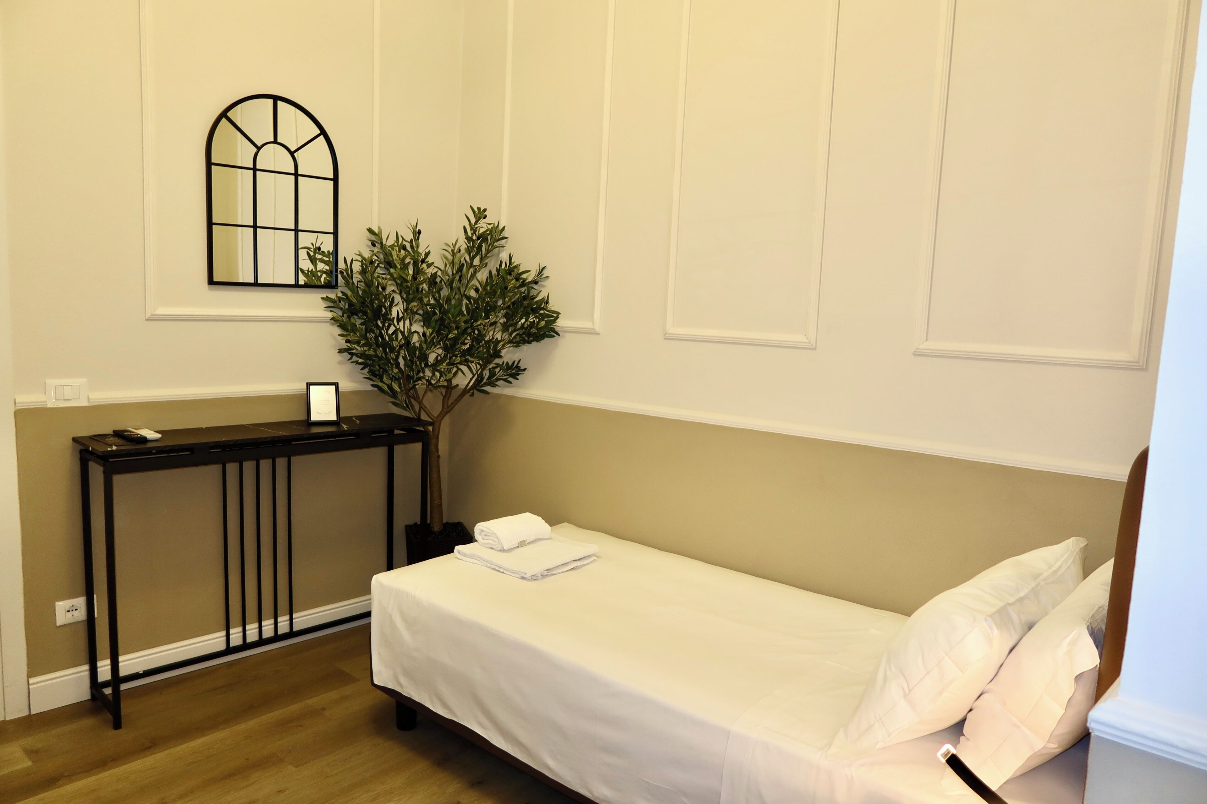A neatly arranged hotel room with a single bed, a small table, a potted plant, and a mirror on the wall.