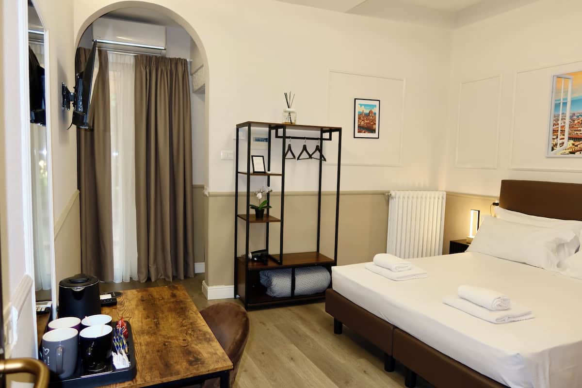 Ett hotellrum med en säng, ett skrivbord med en kaffebryggare, en stol, en klädställning och ett fönster med gardiner.