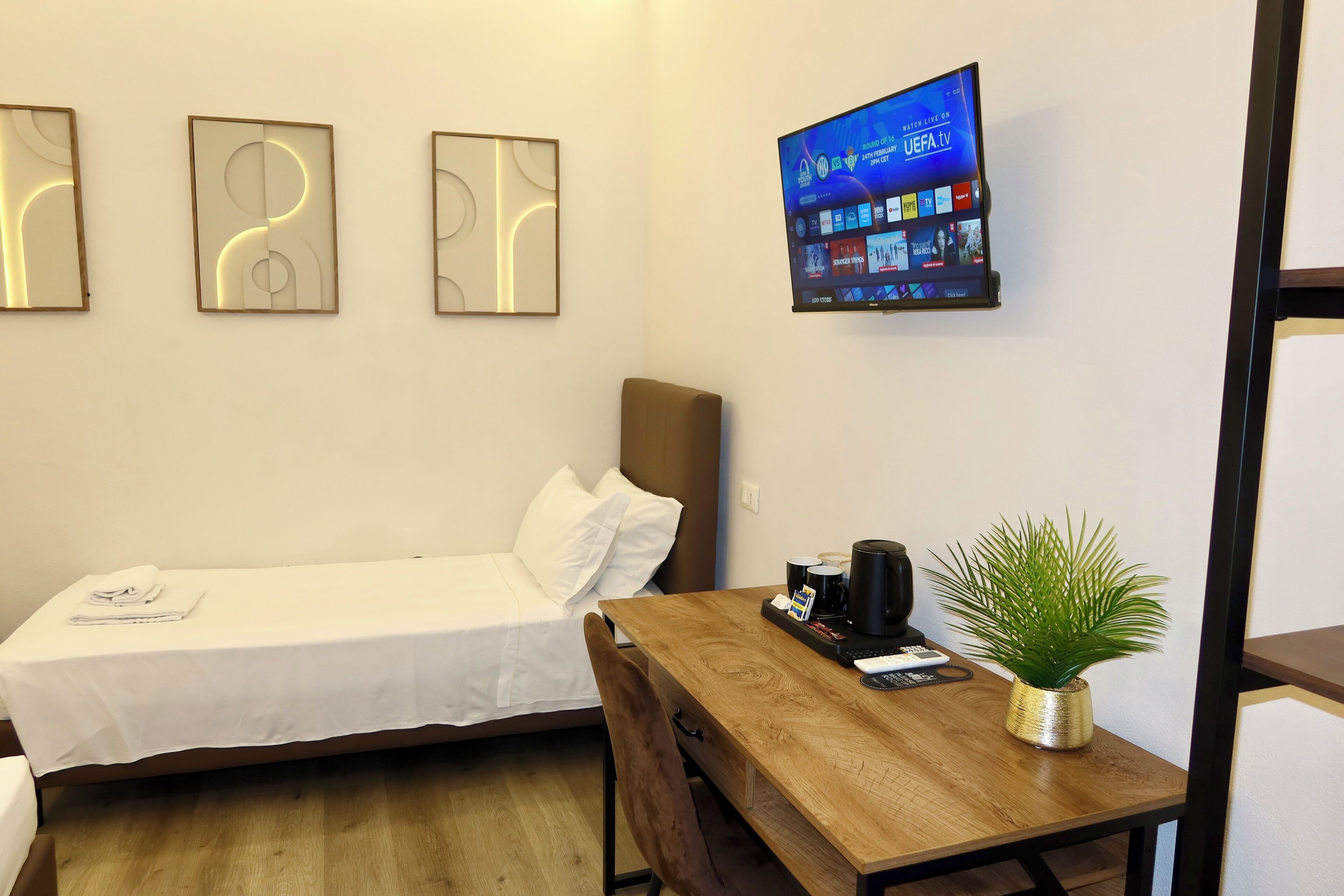 A hotel room with a bed, a wooden table with a plant, a coffee maker, and a TV displaying UEFA TV.