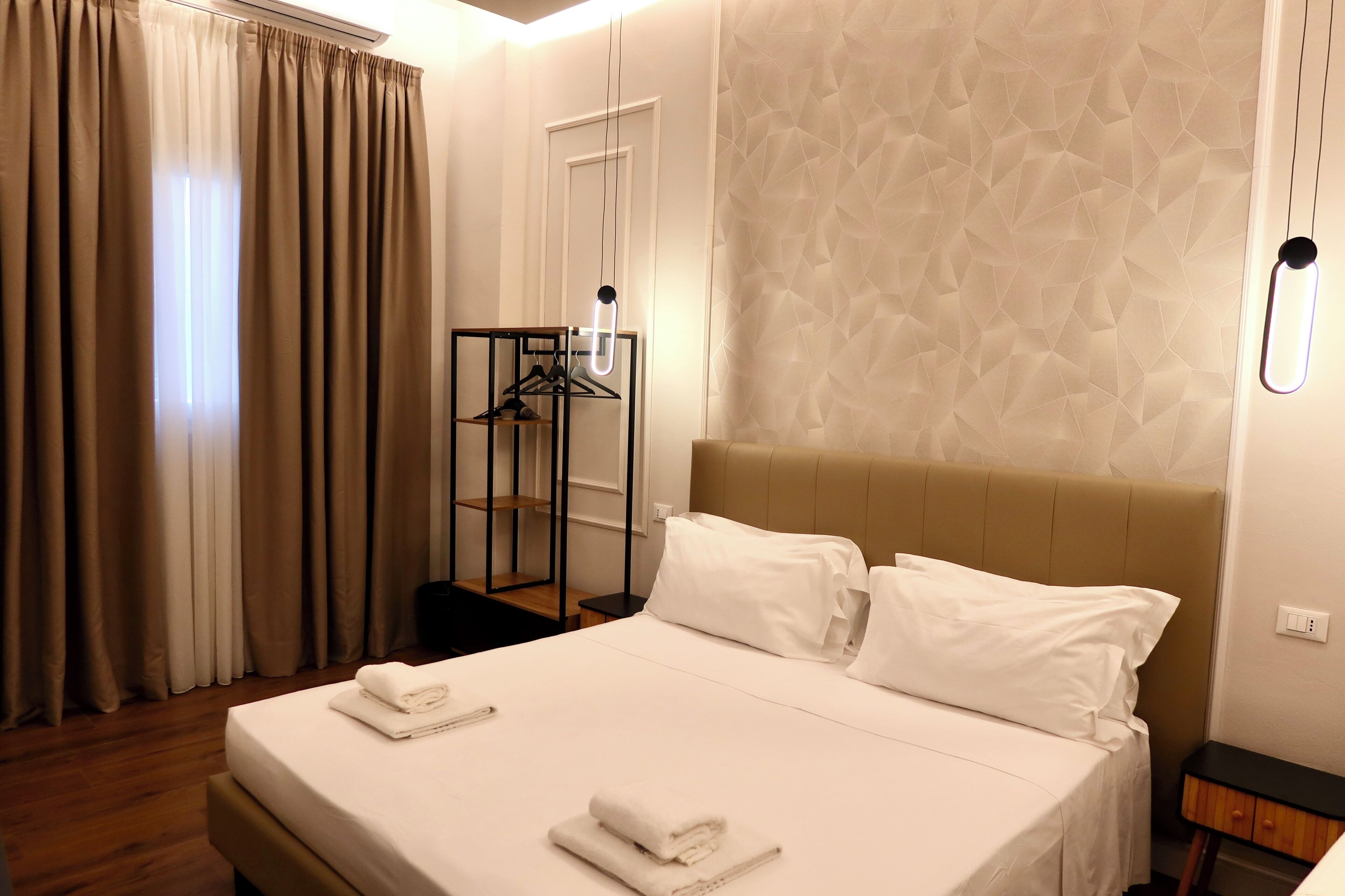 A hotel room with a neatly made bed, a hanging light fixture, a coat rack, and a geometric-patterned wall.