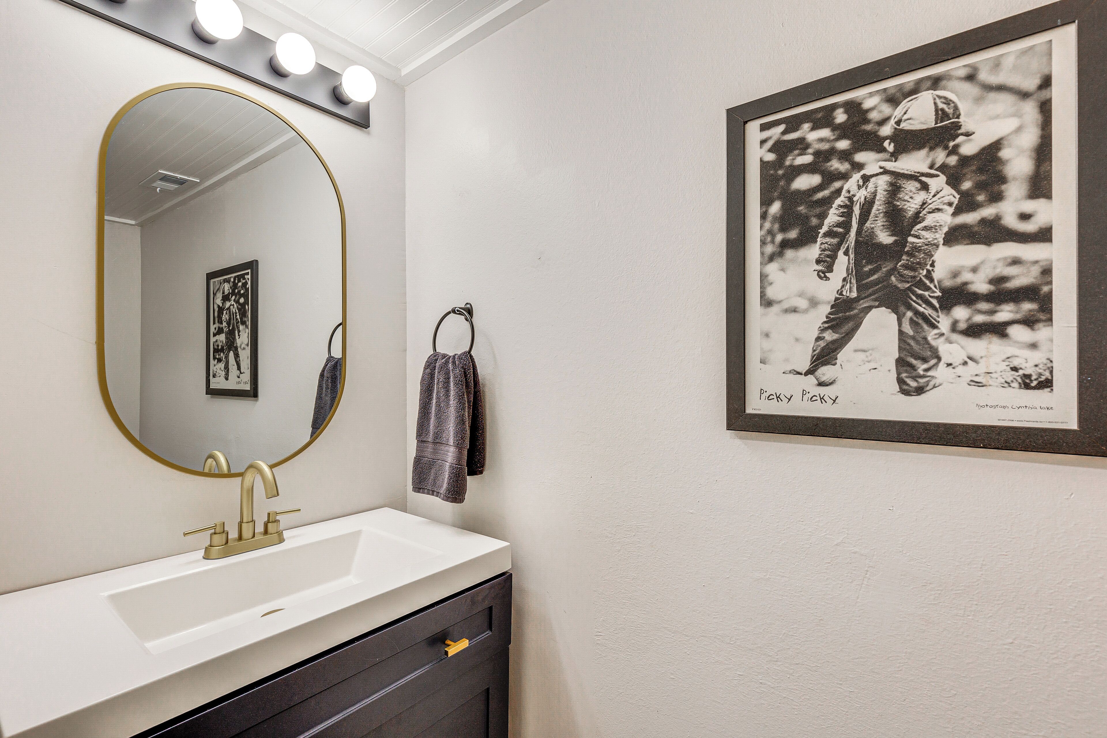 A bathroom with a white sink, gold faucet, and a large mirror with lights. There is a framed black and white photo on the wall.