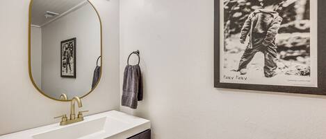 A bathroom with a white sink, gold faucet, and a large mirror with lights. There is a framed black and white photo on the wall.
