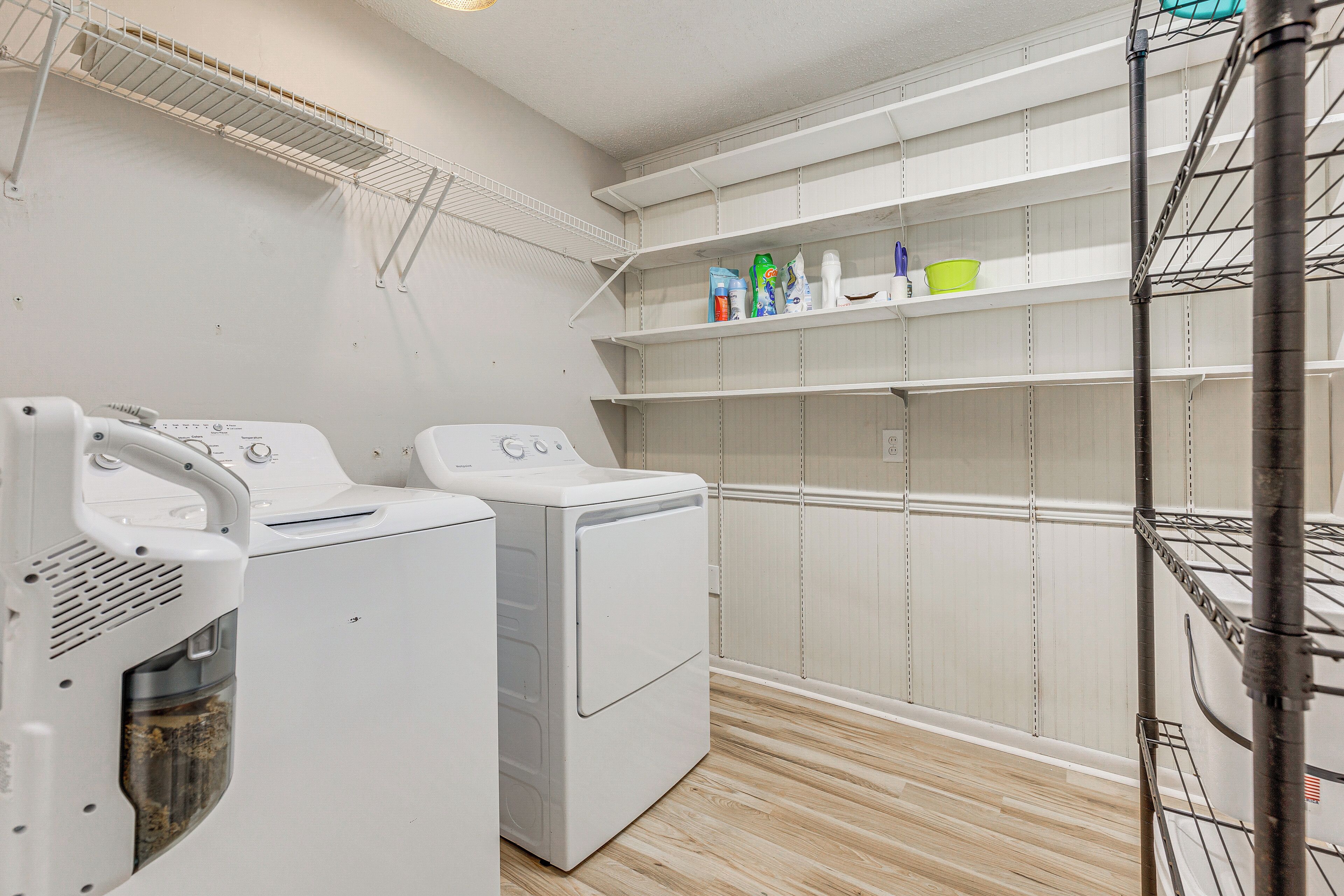 A laundry area with two white washing machines, a metal drying rack, and shelves with cleaning supplies.