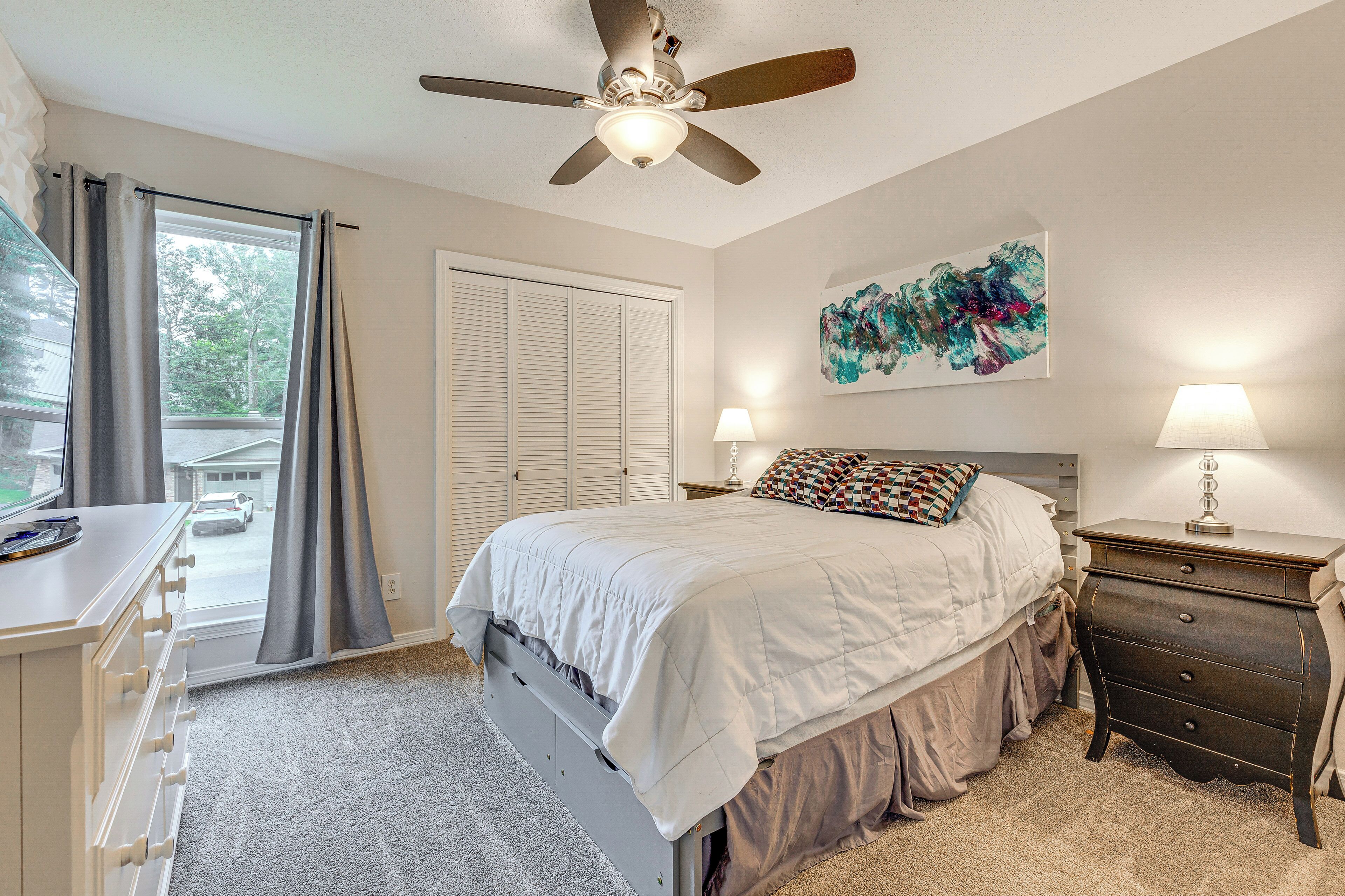 Une chambre à coucher comprenant un lit, une commode, une table de chevet, un ventilateur de plafond, une fenêtre avec des rideaux et un tableau abstrait accroché au mur.