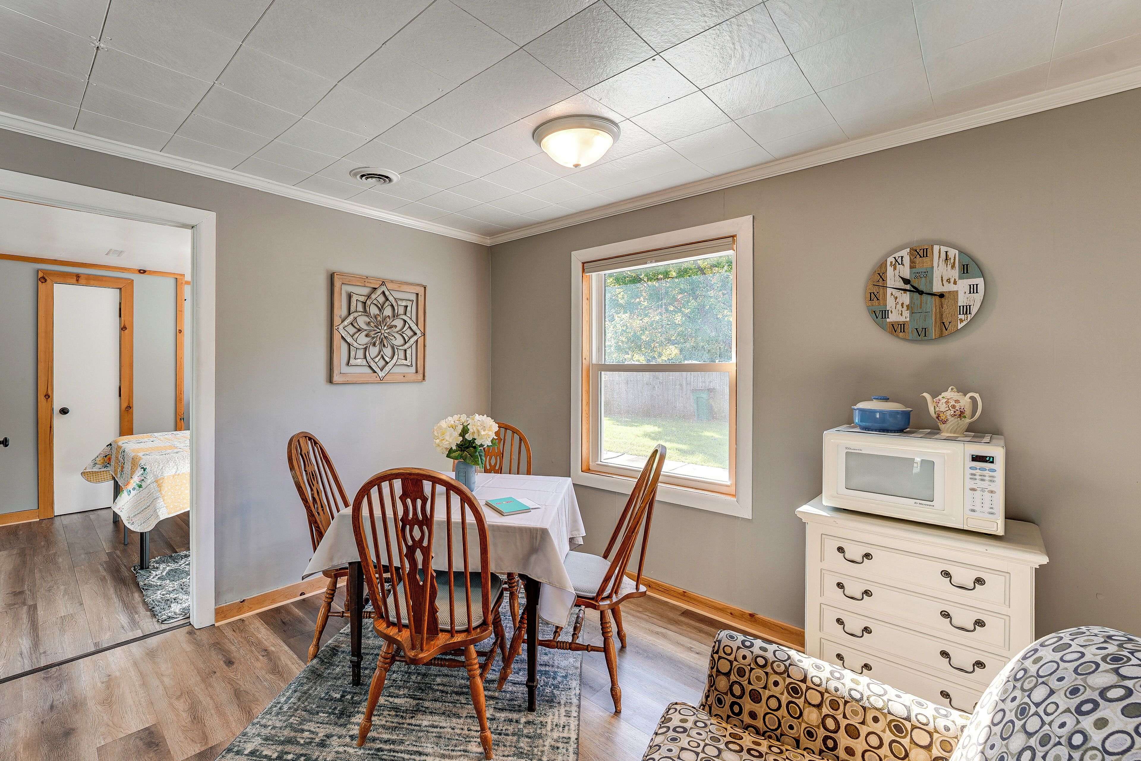 A dining area with a table, chairs, a clock, a microwave, and a window with a view of greenery.