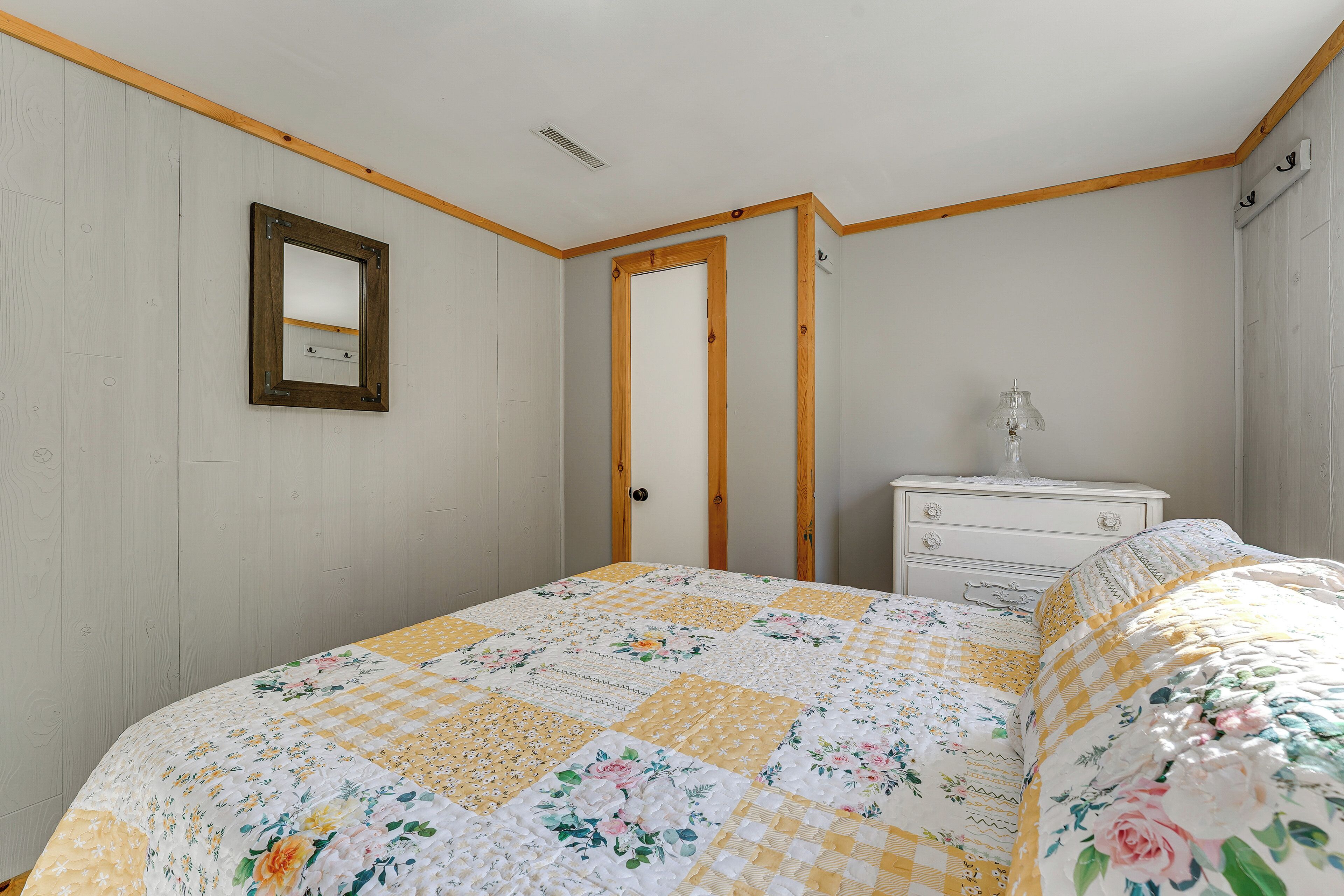 A bedroom with a floral quilt, a wooden headboard, a white dresser, and a mirror on the wall.