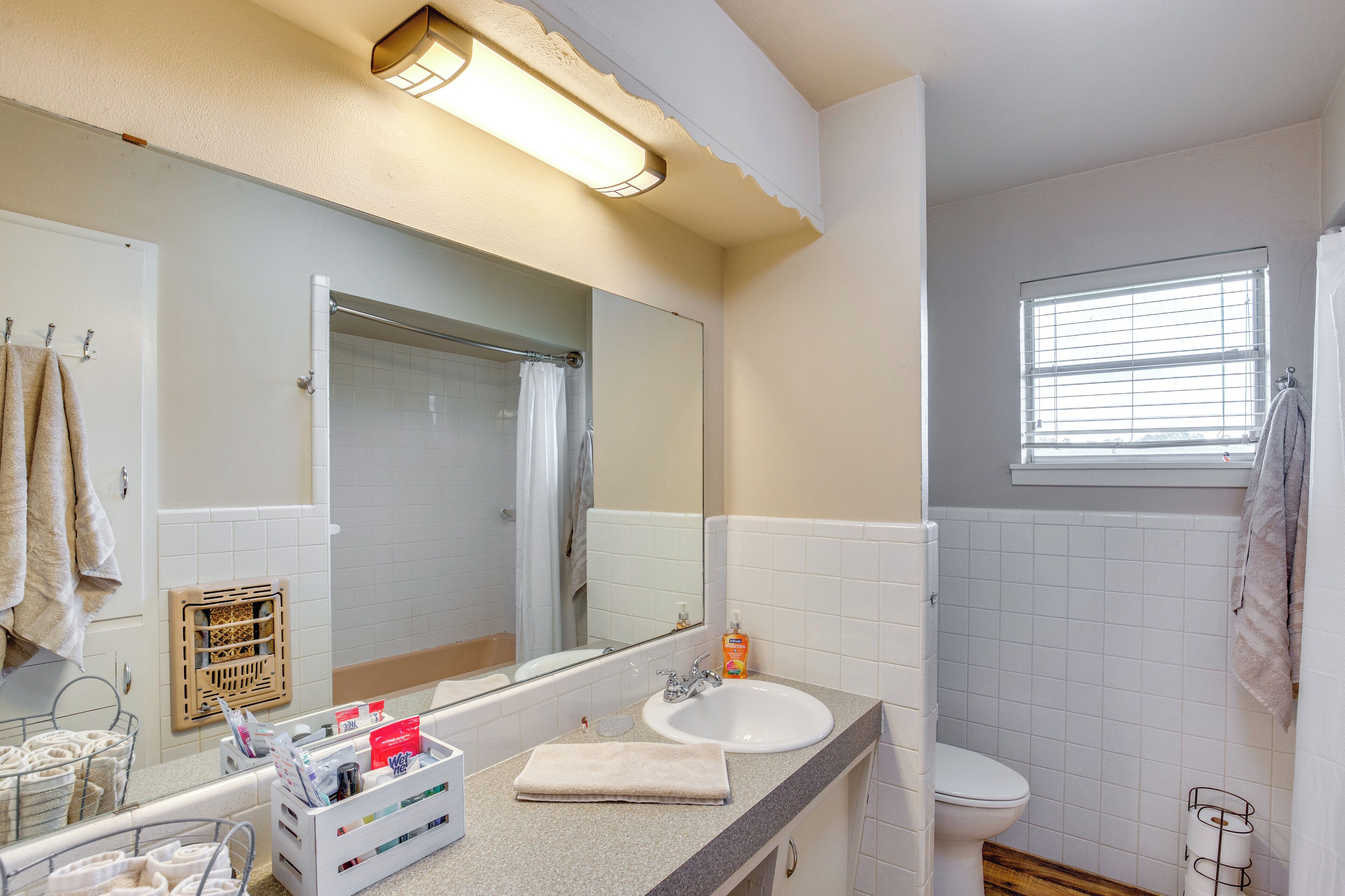 A bathroom with a sink, mirror, and toilet. There is a window with blinds, a towel rack, and a shower curtain.