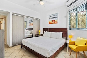 A hotel room with a bed, a chair, a nightstand, a lamp, and a window with blinds. - Aruba Private Pool Oasis: 5BR near Amazing Beaches (Noord)