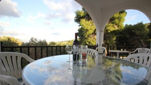 Une table ronde en verre, entourée de chaises blanches, une bouteille de vin et des verres, le tout sur une terrasse extérieure offrant une vue sur les arbres et les collines.