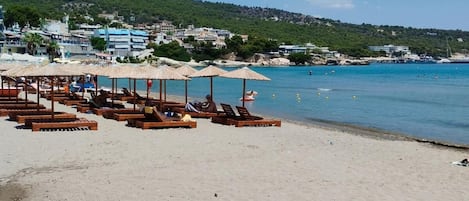 Una playa con sillones de madera y sombrillas, un cielo azul despejado y edificios al fondo.