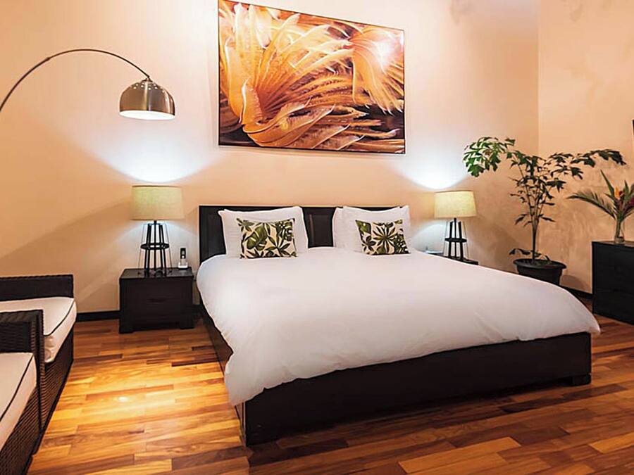 A modern hotel room with a large bed, two bedside tables with lamps, a floor lamp, a sofa, and a large painting on the wall.