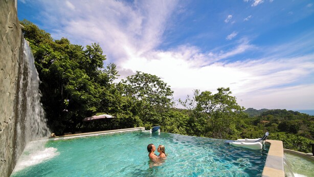 A clear pool with a waterfall feature, surrounded by lush greenery and a view of distant hills. - Gaia Collection Hotels - Adults only (Quepos)