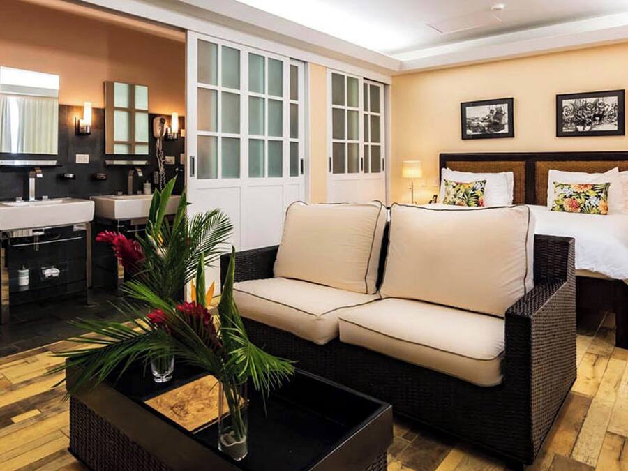 A modern hotel room with a sofa, a small coffee table with a plant, a bed with two pillows, a nightstand, and a bathroom with a sink and mirror.