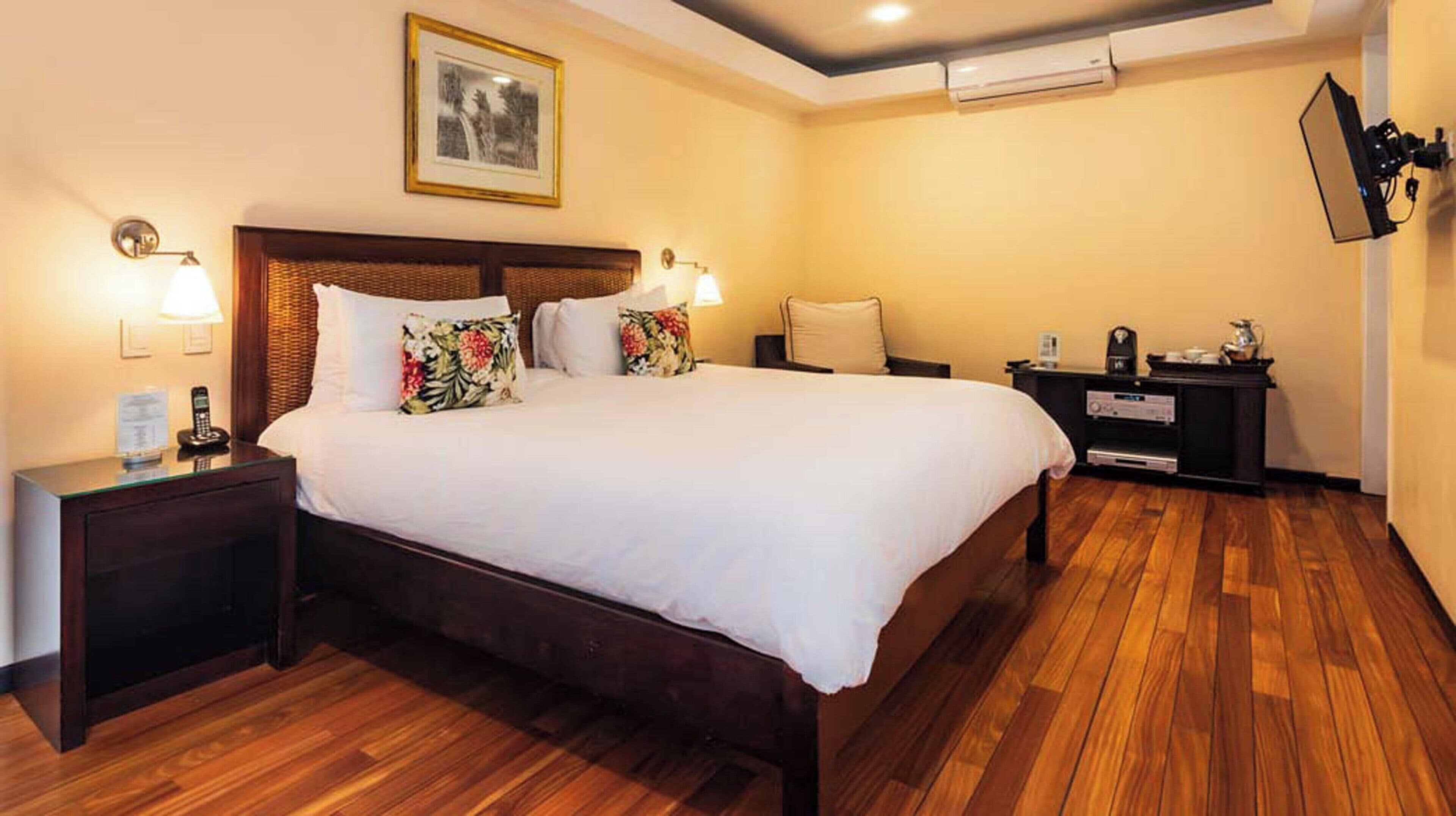 Una camera d'albergo con un letto grande, pavimento in legno, un comodino con un telefono, un tavolino e un condizionatore d'aria a parete.