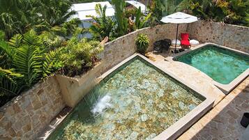 En swimmingpool med en stenvæg og frodig beplantning, et opholdsområde med stole og en parasol, samt et lille vandfald.