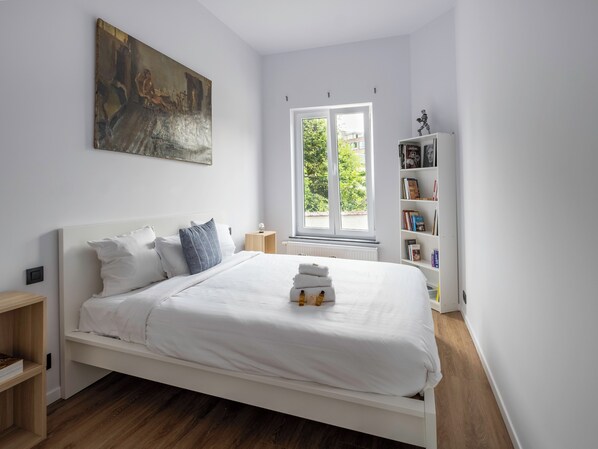 Un dormitorio moderno con una cama blanca, un cuadro en la pared, una estantería y una ventana con vistas a zonas verdes.
