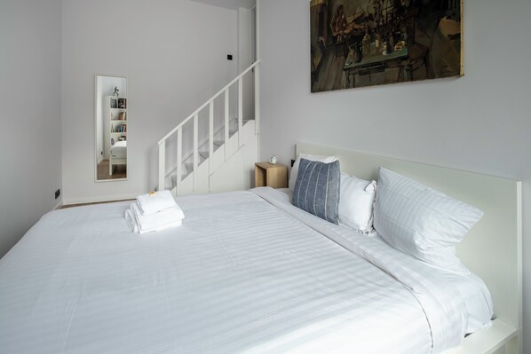 A neatly made bed with white linens and a folded towel, a wooden nightstand with a lamp, a painting on the wall, and a white staircase. - Brussels Toison D'or (Brussels)