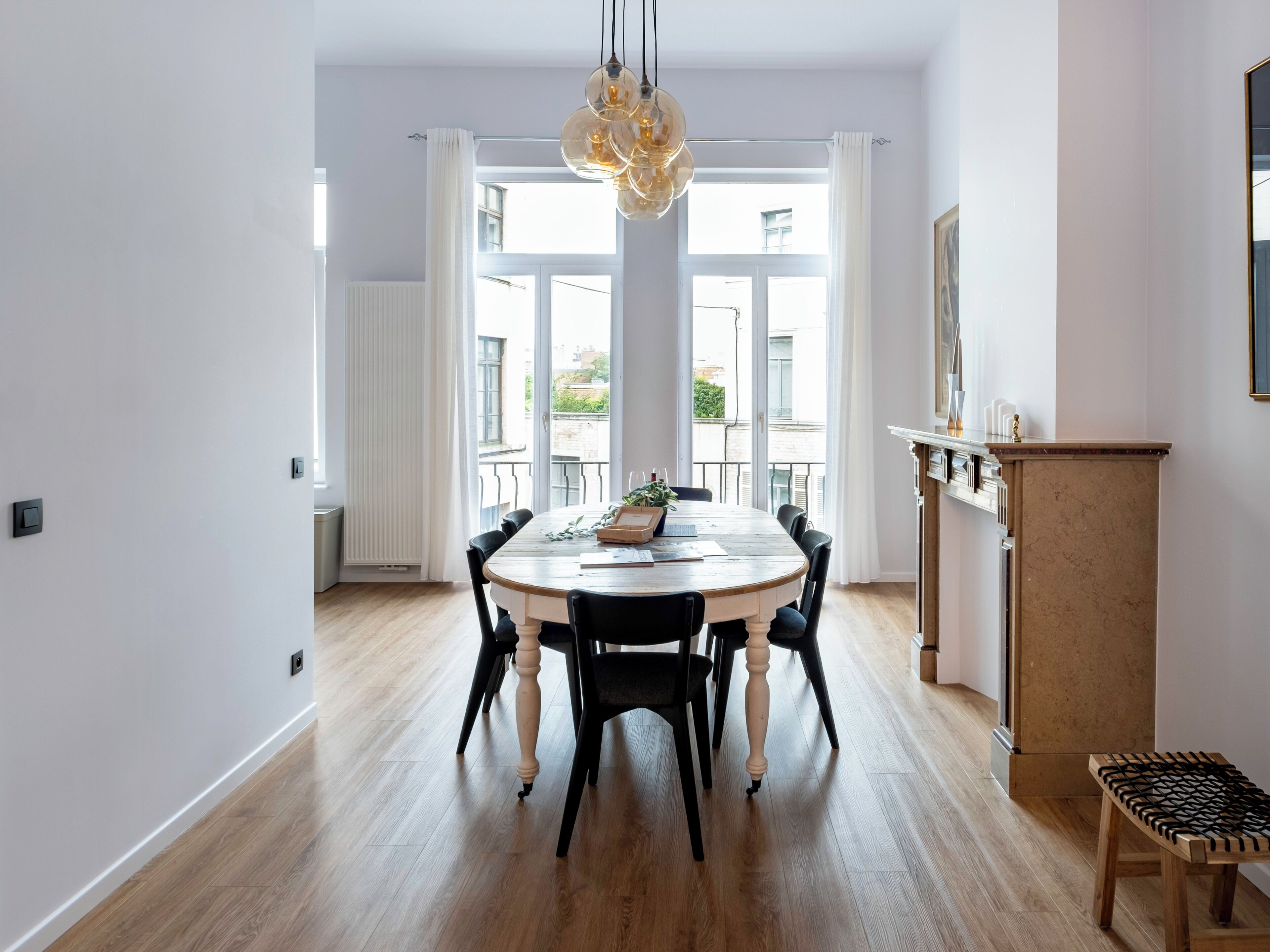 Un espace repas avec une table ronde, des chaises noires, un plancher en bois, un luminaire suspendu et une vue sur l’extérieur grâce à de grandes fenêtres.