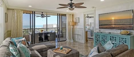 Een ruime woonkamer met een grote bank, een salontafel en uitzicht op de oceaan. Er is een plafondventilator, een televisie en een eethoek met uitzicht op het balkon.