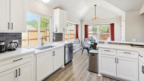 A modern kitchen with white cabinets, a dishwasher, and a coffee maker. There is a dining area with a table and chairs, and a window with curtains.