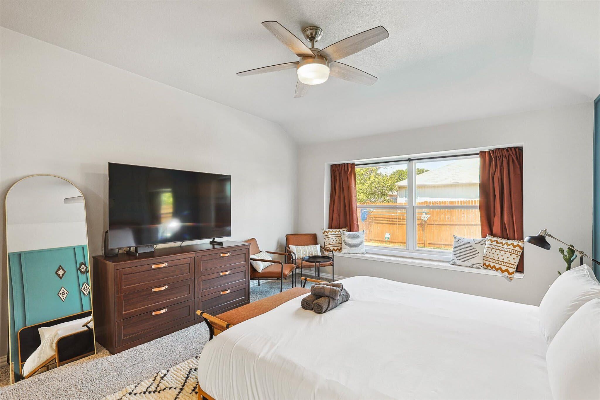 Une chambre d’hôtel équipée d’un lit, d’une télévision, d’une commode, d’une chaise, d’une fenêtre avec des rideaux et d’un ventilateur de plafond.