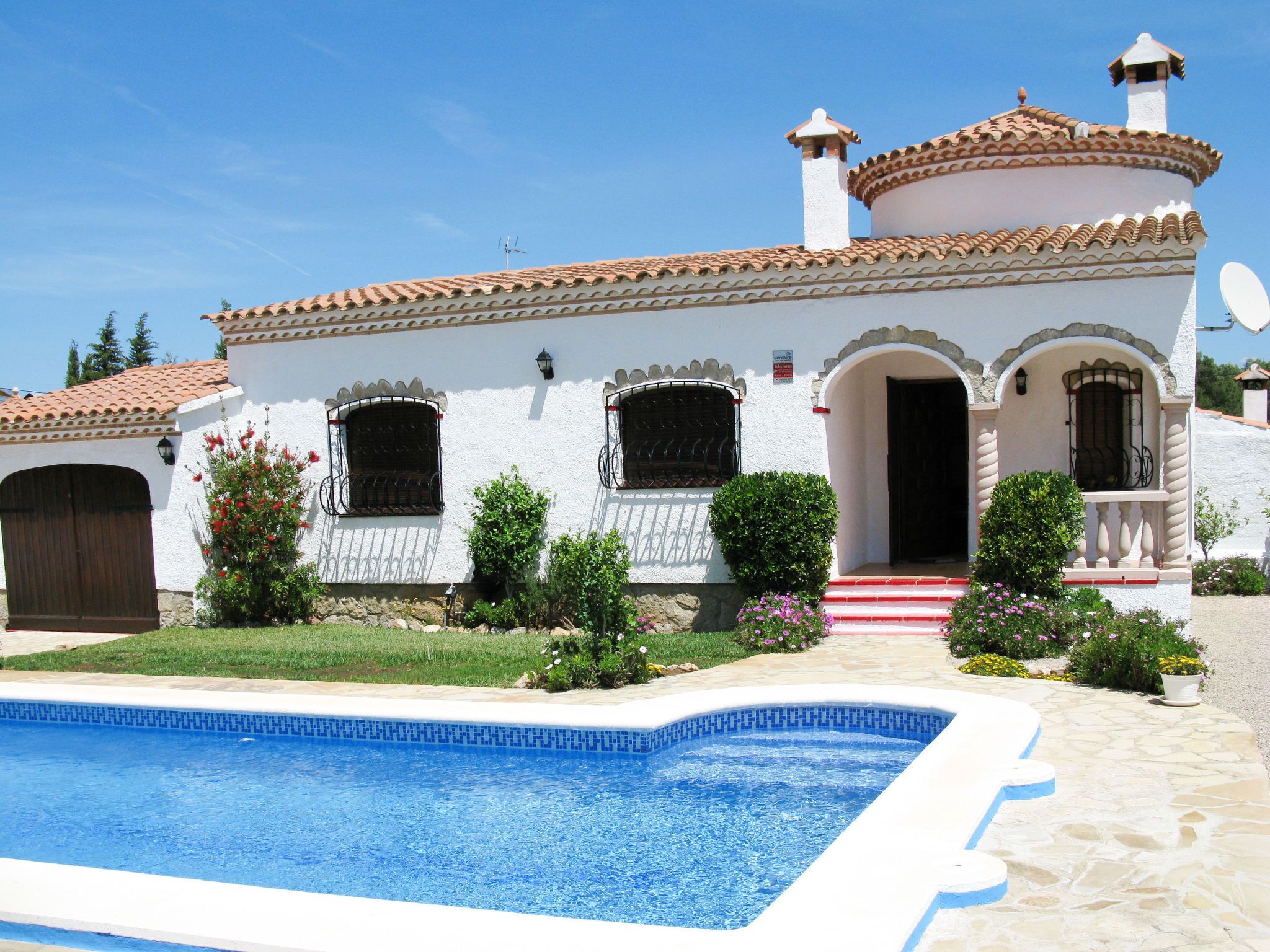 Une villa blanche dotée d’une piscine, d’un toit en tuiles et d’un jardin agrémenté de fleurs et d’arbustes.