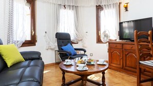 Un salon avec un canapé en cuir noir, une table à manger en bois sur laquelle se trouvent des tasses et un gâteau, un fauteuil noir avec un coussin bleu, et un téléviseur posé sur un meuble en bois.