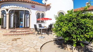Un bâtiment blanc avec un toit en tuiles, un patio en pierre avec des sièges d’extérieur et un arbuste vert.