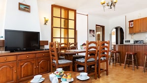 Un coin repas avec une table en bois, dressée pour quatre personnes, entourée de chaises en bois. Un téléviseur est posé sur un meuble en bois, à gauche.