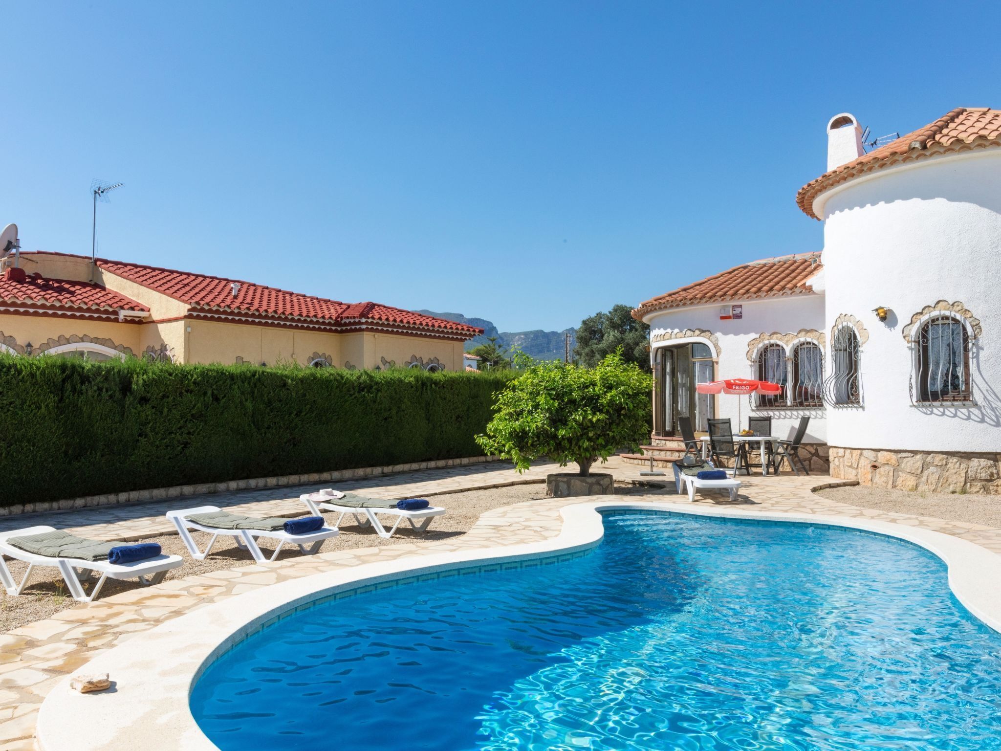 A swimming pool with lounge chairs and a small table, surrounded by a white building with a red-tiled roof.