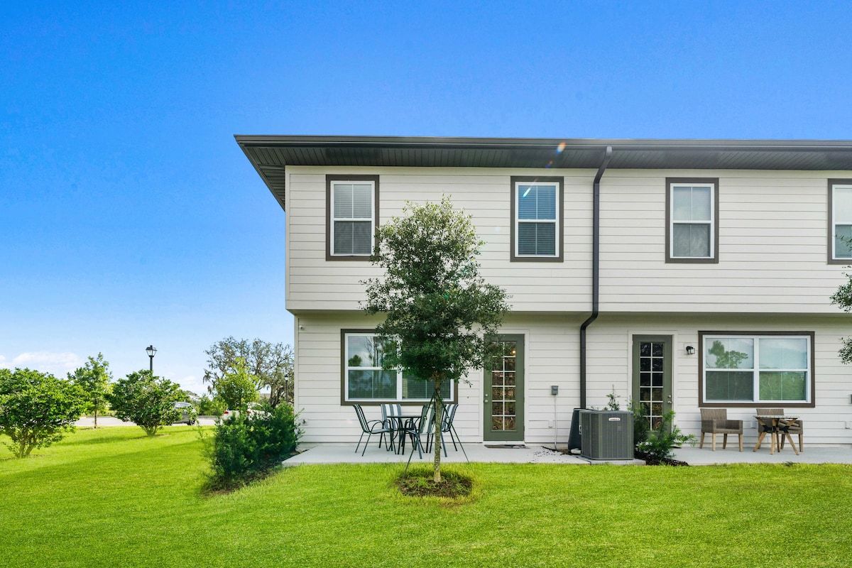 A two-story residential building with a front lawn, a small tree, and outdoor seating.