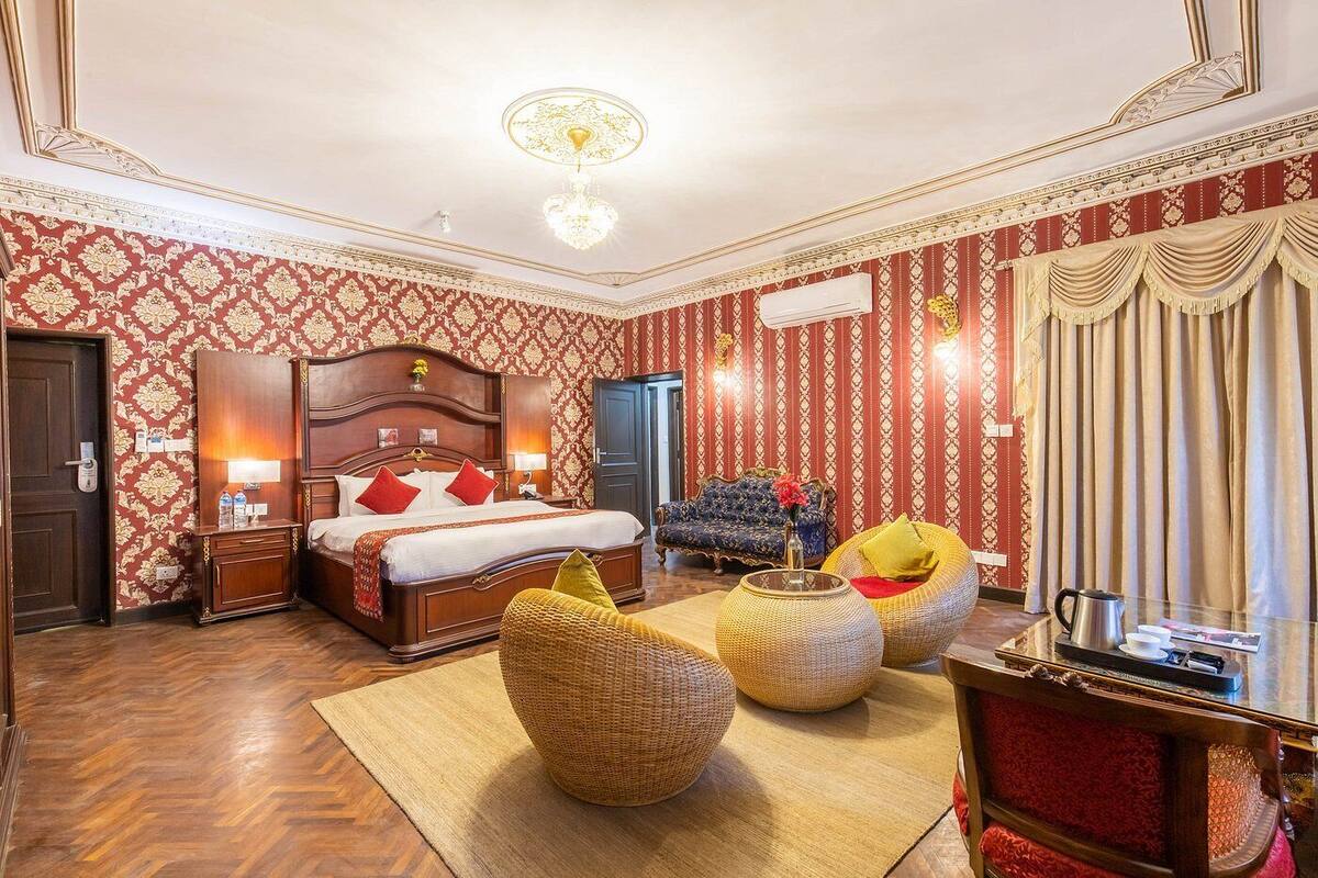 Amplia habitación de hotel con una cama grande, una zona de estar con sofá y sillones, una mesita con hervidor y un espejo.
