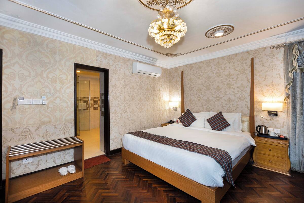 Habitación de hotel con una cama grande, cabecero de madera, una mesita de noche con lámpara, una pequeña mesa con hervidor y un zapatero.