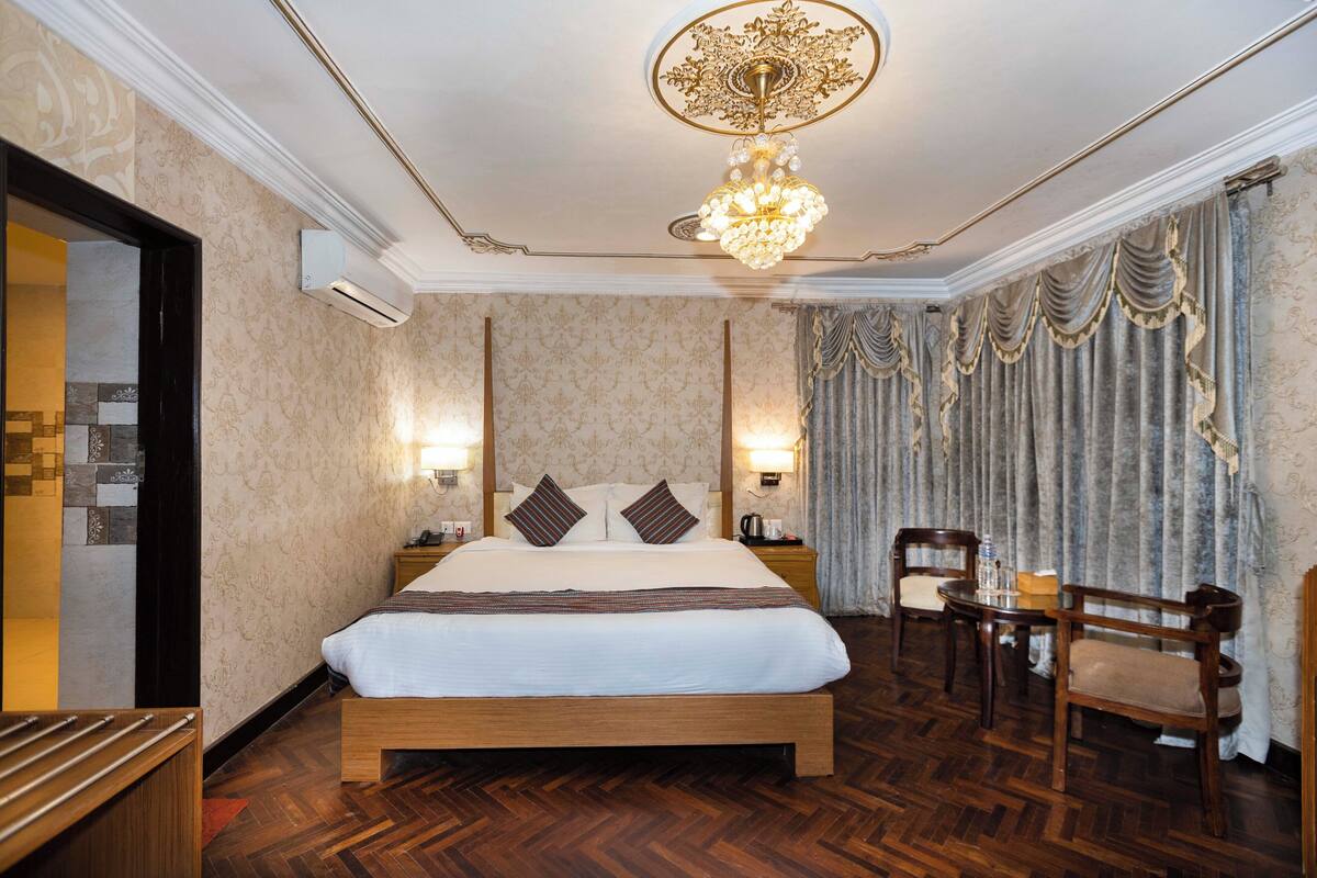 Una habitación de hotel con una cama grande, cabecera de madera, una araña, un escritorio con silla y un ventanal con cortinas.