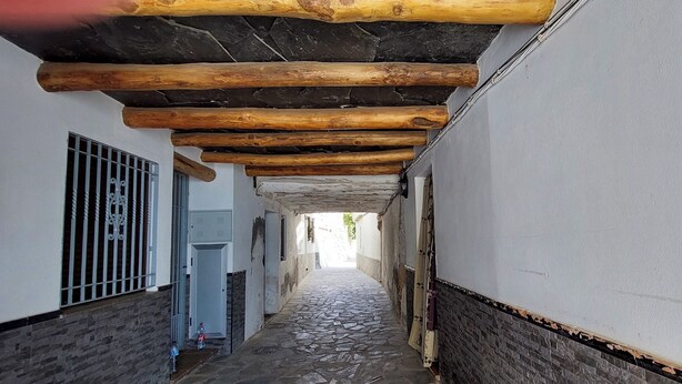 Un couloir étroit, pavé de pierres, avec des poutres en bois au plafond, des murs blancs et un portail en métal sur le côté gauche.