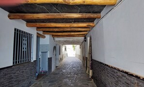 Ein schmaler, mit Kopfsteinpflaster belegter Gang mit Holzbalken an der Decke, weißen Wänden und einem Metallgitter links.