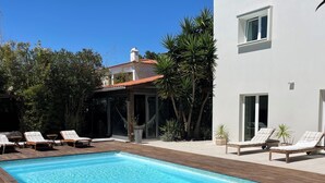A rectangular swimming pool with wooden decking, surrounded by lounge chairs and a white building with a red roof.